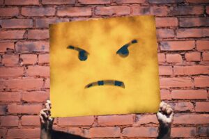 Wütend dreinschauendes Frowny (wie in "Smiley") auf einen Karton gemalt und von zwei Händen hochgehalten.