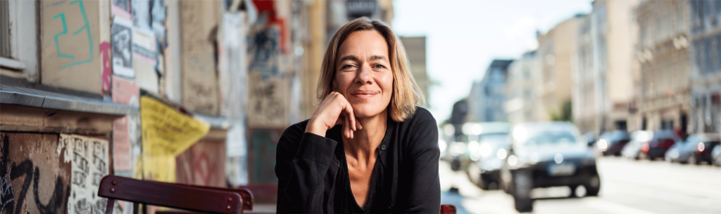 Jule sitzt an einem Gastro-Tisch neben einer stark plakatierten Hauswand. Sie lächelt in Richtung der Kamera.